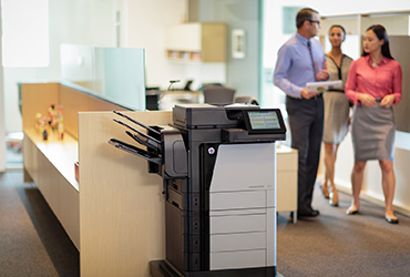 Coworkers walk by an HP Laserjet MFP 630 printer in an office.